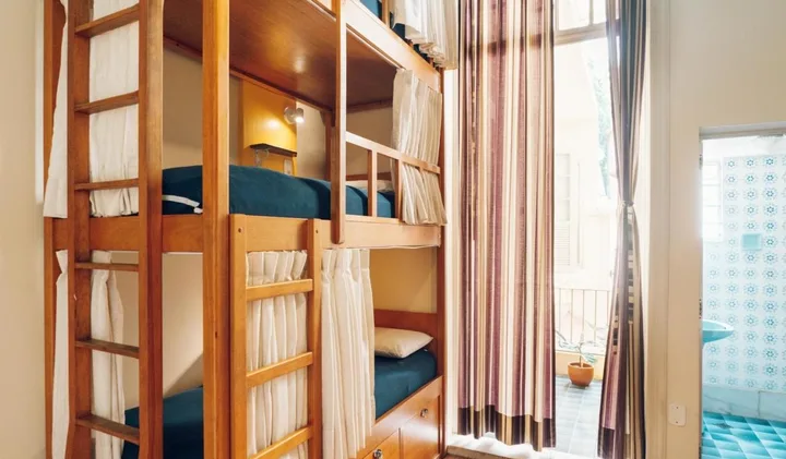 Wooden bunk beds with privacy curtains in a bright, well-lit hostel dormitory room.