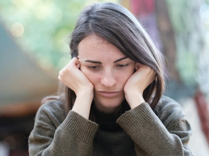 Young woman sitting outdoors with head in hands, looking downward with a sad, worried expression.