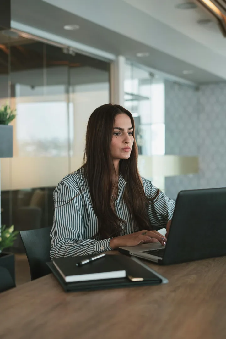 Professional woman in striped shirt working focused at a laptop in a modern office setting.