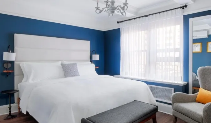 Modern hotel room with bold blue walls, white upholstered headboard, chandelier, and armchair.