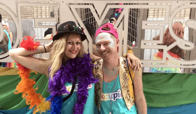Two festively dressed people posing together at a colorful Pride event with feather boas and glitter.