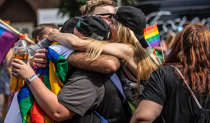 Group of people hugging at a Pride event, with rainbow flags and festive atmosphere outdoors.