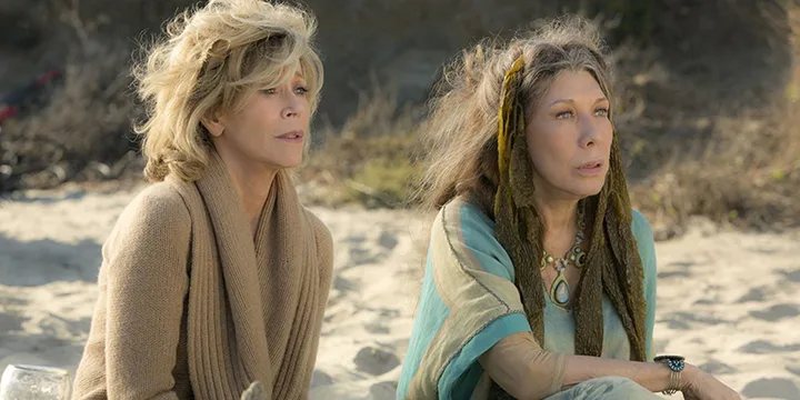 Two older women sitting on a sandy beach, windswept hair, in a TV drama scene.