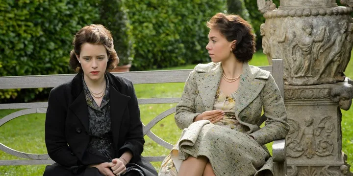 Two women in 1940s period costumes sit on a garden bench in a dramatic TV scene.
