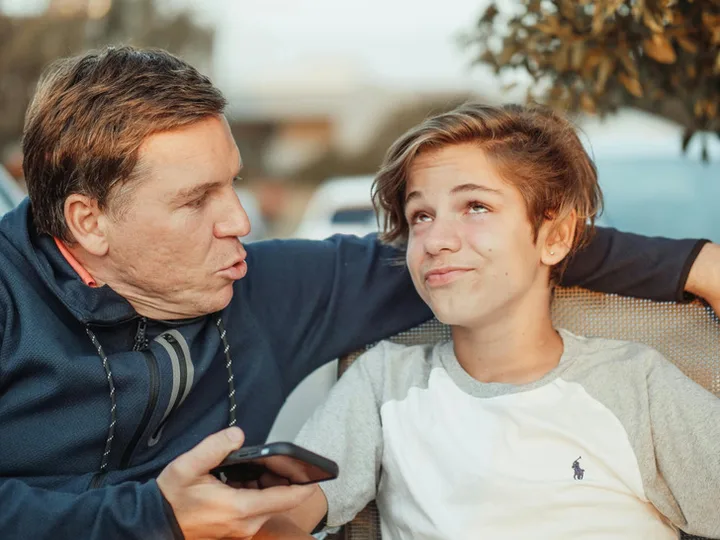 Middle-aged man with arm around a teenage boy, both smiling in a warm outdoor moment.