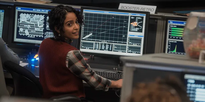 Woman smiles at camera while seated at a mission control console with multiple technical screens.