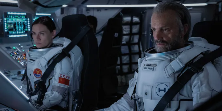 Two actors in white space suits sit inside a detailed spacecraft cockpit with glowing instrument panels.