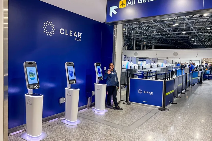 CLEAR Plus biometric kiosks with a smiling staff member waving at an airport security area.