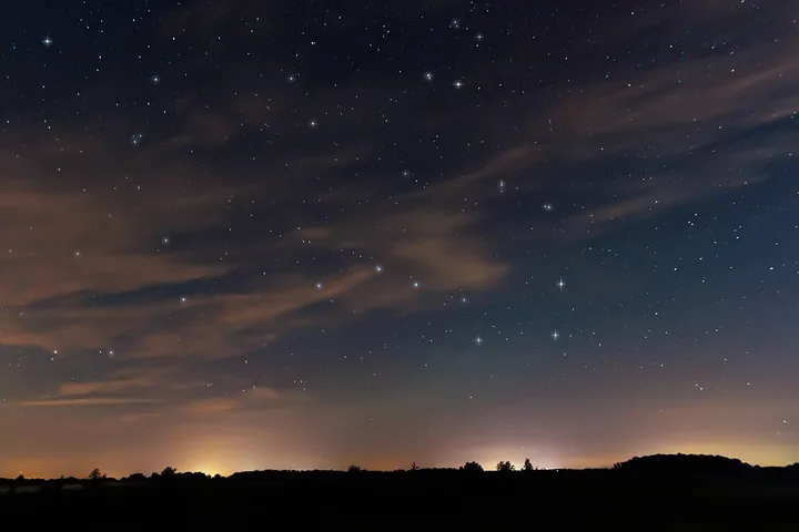 Wide-angle night sky photo showing stars and wispy clouds above a dark rural horizon with distant city glow.