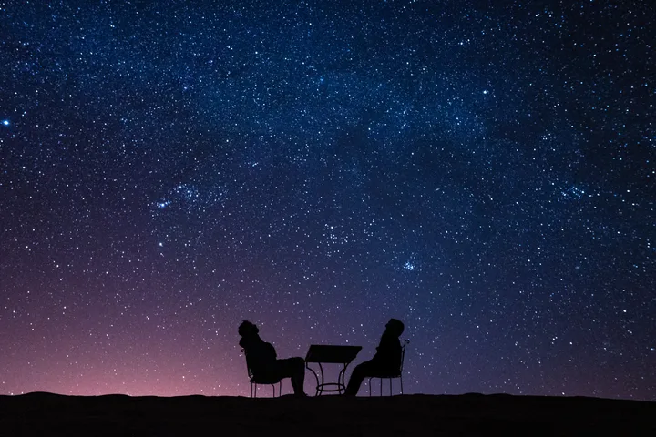 Two silhouetted figures sitting at a table under a vast, star-filled purple night sky.