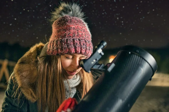 Young woman in winter hat peering through a telescope eyepiece outdoors at night.