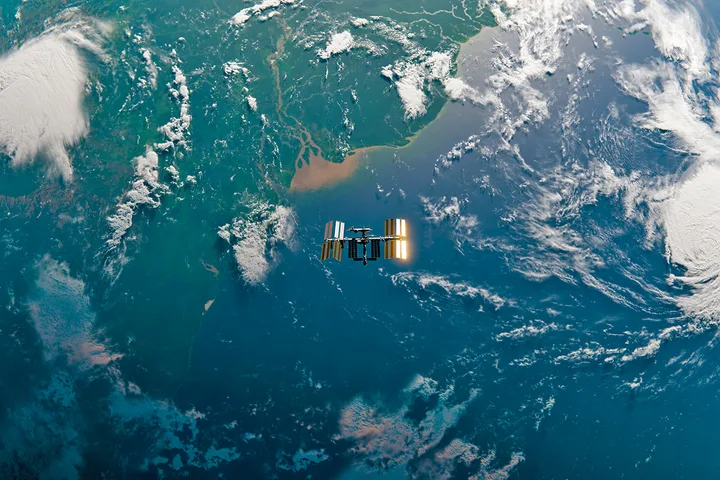 International Space Station photographed from above against Earth's curved surface with ocean and clouds.