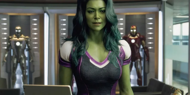 She-Hulk character in superhero suit stands confidently in a room with Iron Man armor displays behind her.