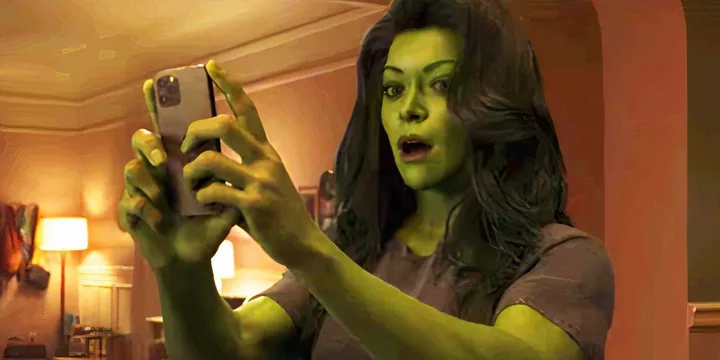 She-Hulk character with green skin looks shocked while holding a smartphone in a warmly lit room.