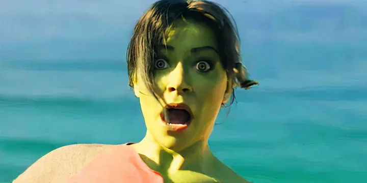 She-Hulk character with wide shocked eyes and open mouth stands in front of turquoise ocean water.