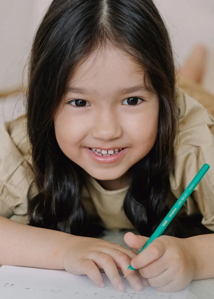 Smiling young girl lying down drawing with a green pencil, looking warmly at the camera.
