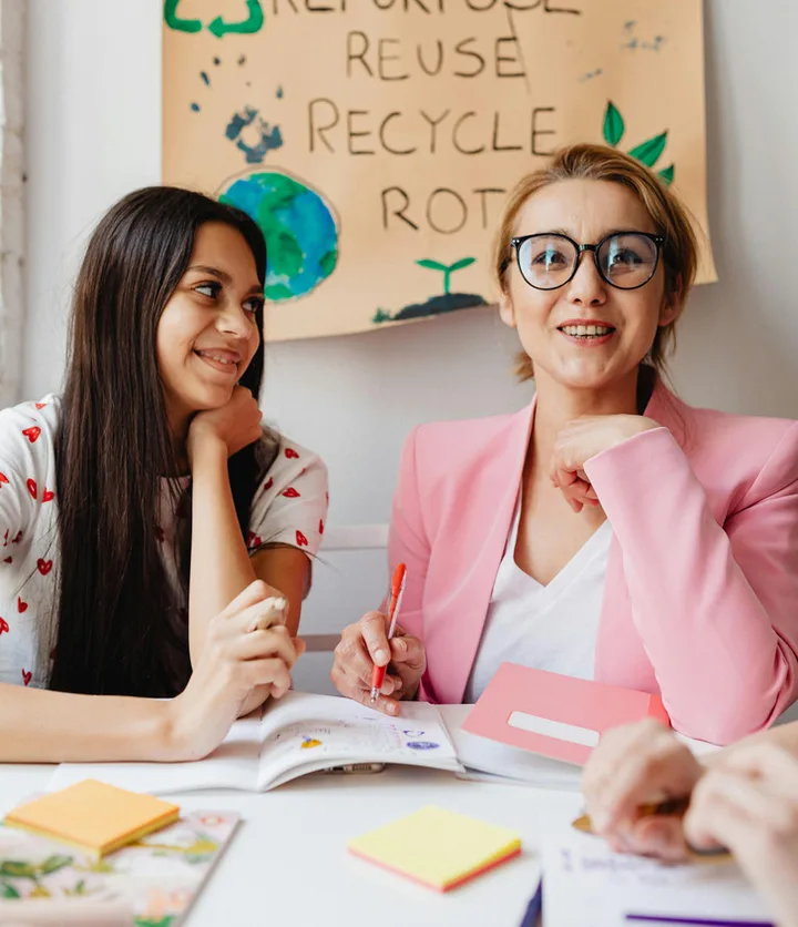 Teacher and teenage student smiling together at a desk in a classroom with recycling poster.