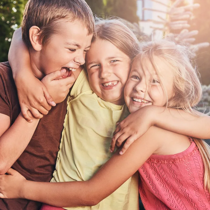 Three laughing children hugging each other joyfully outdoors in warm sunlight.