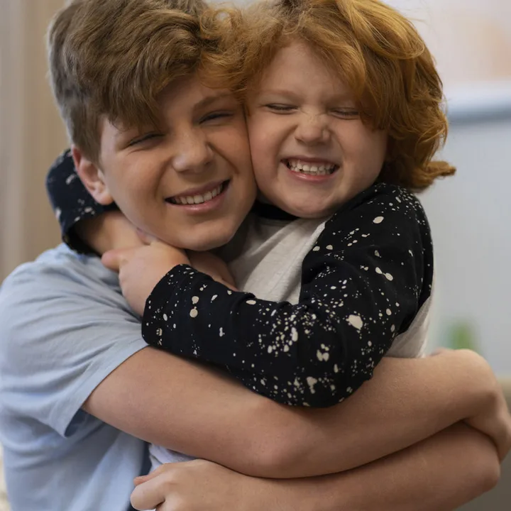 Two boys laughing and hugging each other tightly indoors with big smiles.