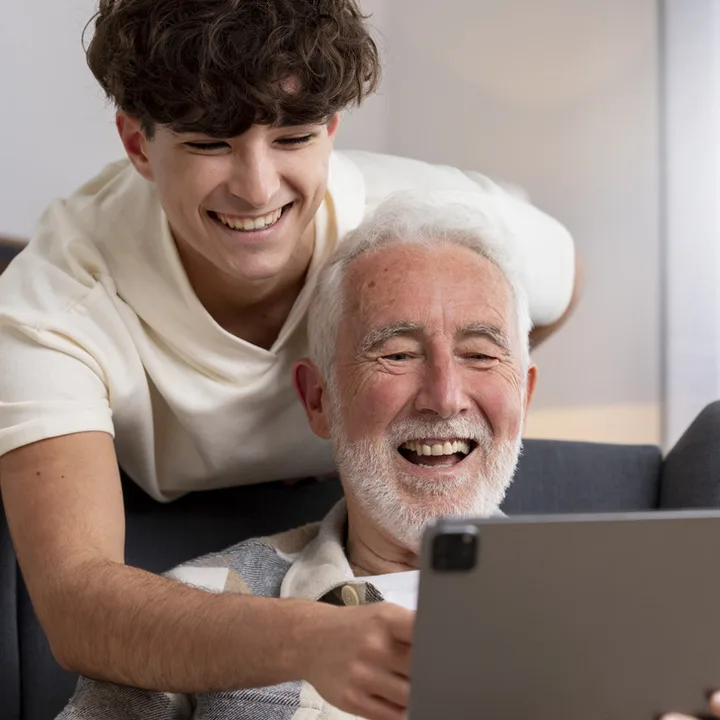 Smiling teenage boy helping an elderly man with a tablet, both laughing indoors.