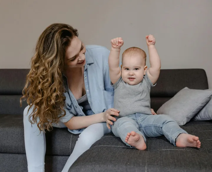 Smiling young woman sitting on a couch beside a cheerful baby with arms raised.