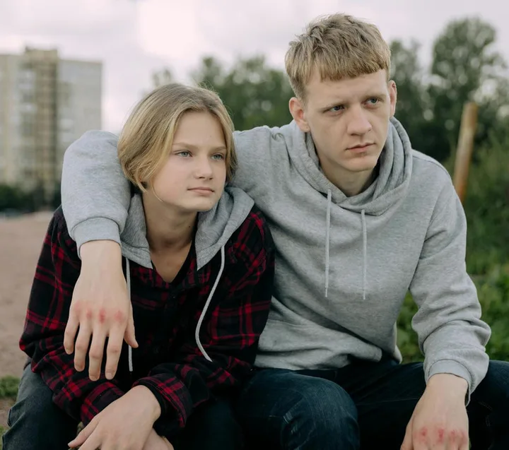 Two teenagers sitting together outdoors, one resting arm around the other, both with serious expressions.
