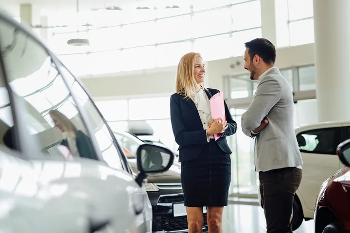 A female car salesperson laughs while talking with a male customer among vehicles in a bright showroom.