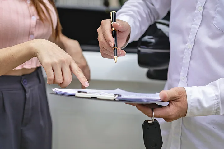 Close-up of a salesman holding a clipboard and car keys while a woman points at paperwork.