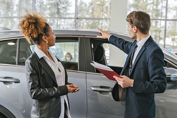 A male salesman in a suit gestures toward a gray car while speaking with a female customer in a dealership.