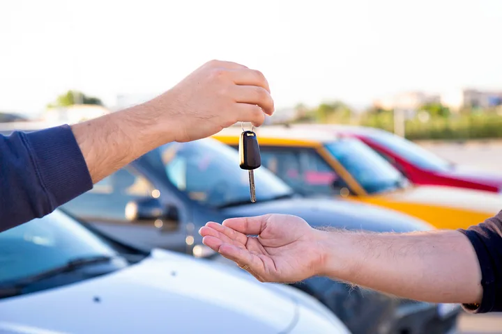 Two hands exchanging a car key in an outdoor car lot with colorful vehicles in the background.
