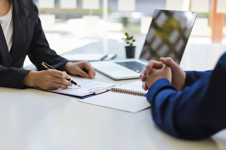 A businesswoman reviews documents with a pen across a desk from a client with a laptop nearby.