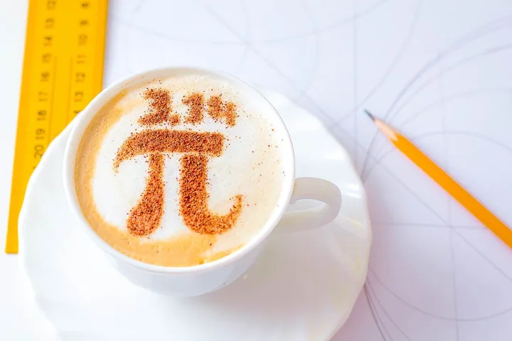A latte with the pi symbol drawn in cinnamon on the foam, beside a ruler and pencil.