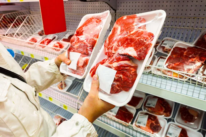 Shopper holds up two packaged meat trays for comparison in a grocery store meat section.