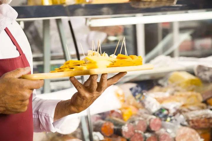 Deli worker in red apron holds a tray of cheese samples on toothpicks near a display case.