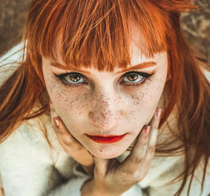 Close-up portrait of a red-haired woman with freckles gazing intensely at the camera, hands on her face.