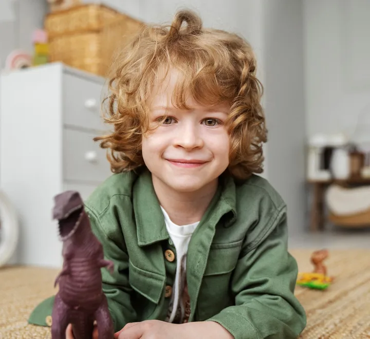 Curly-haired young boy smiling while lying on the floor holding a toy dinosaur in a playroom.
