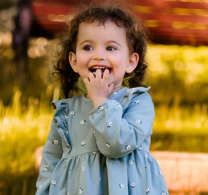 Toddler girl in a blue floral dress laughing joyfully outdoors in warm golden sunlight.