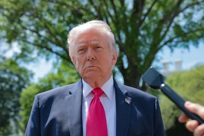 Man in blue suit and red tie speaking to reporters outdoors near trees.