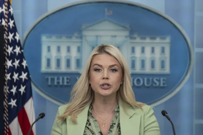 Blonde woman in green blazer speaking at White House briefing room podium with US flag visible.