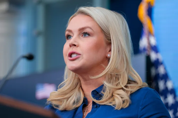 Close-up of blonde woman speaking passionately at a podium with microphones and US flag in background.