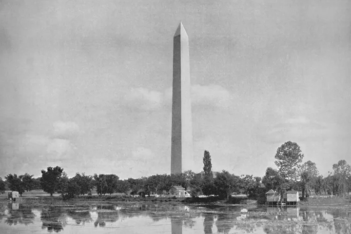 Vintage black-and-white photo of Washington Monument reflected in surrounding wetlands with small structures nearby.