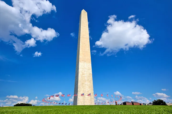 Bright daytime photo of Washington Monument towering against blue sky, surrounded by American flags on green lawn.