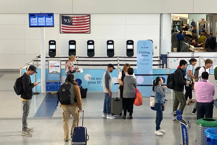 Travelers with luggage queue at a CLEAR security enrollment area inside a busy airport terminal.