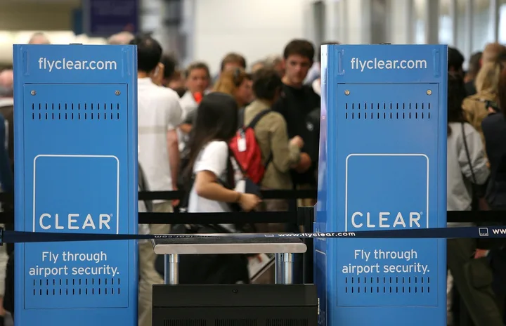 Two blue CLEAR airport security kiosks with 'Fly through airport security' signage, travelers in background.