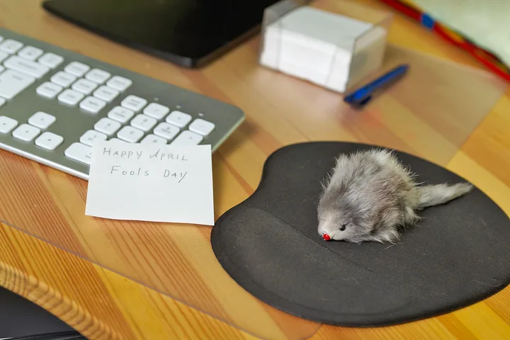 Office desk with a fake toy mouse on a mousepad and a 'Happy April Fools Day' note.