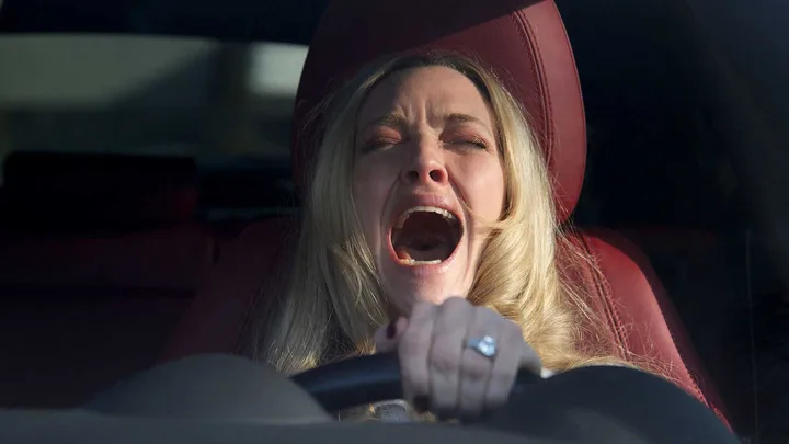 Blonde woman screaming in anguish while gripping a steering wheel inside a car.
