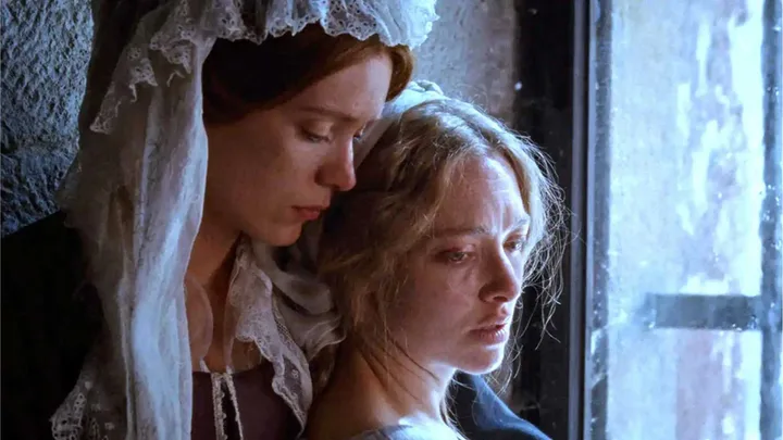 Two women in period costumes leaning together near a window in an intimate, melancholy film scene.