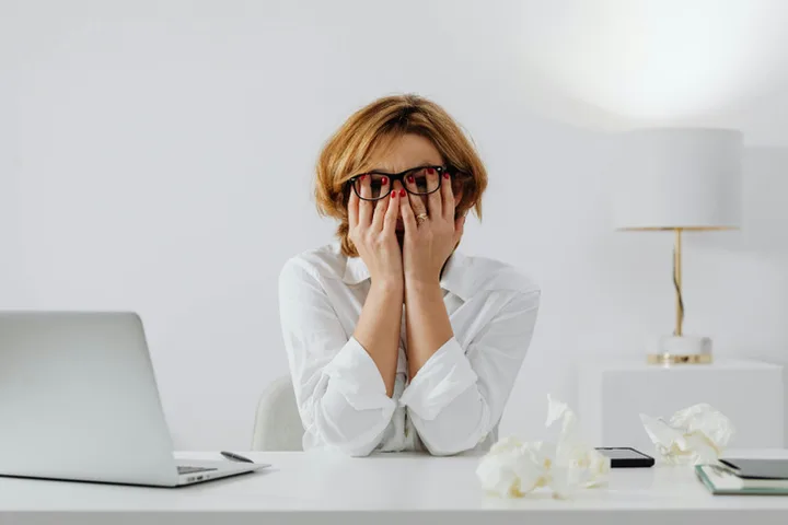 Distressed woman covering her face with hands at a desk, tissues nearby and laptop open, office setting.