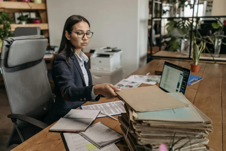 Woman in blazer reviewing a large stack of folders and documents at a cluttered office desk.