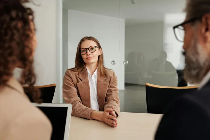 Woman in glasses sitting calmly across a table from two people in an interview or formal meeting setting.
